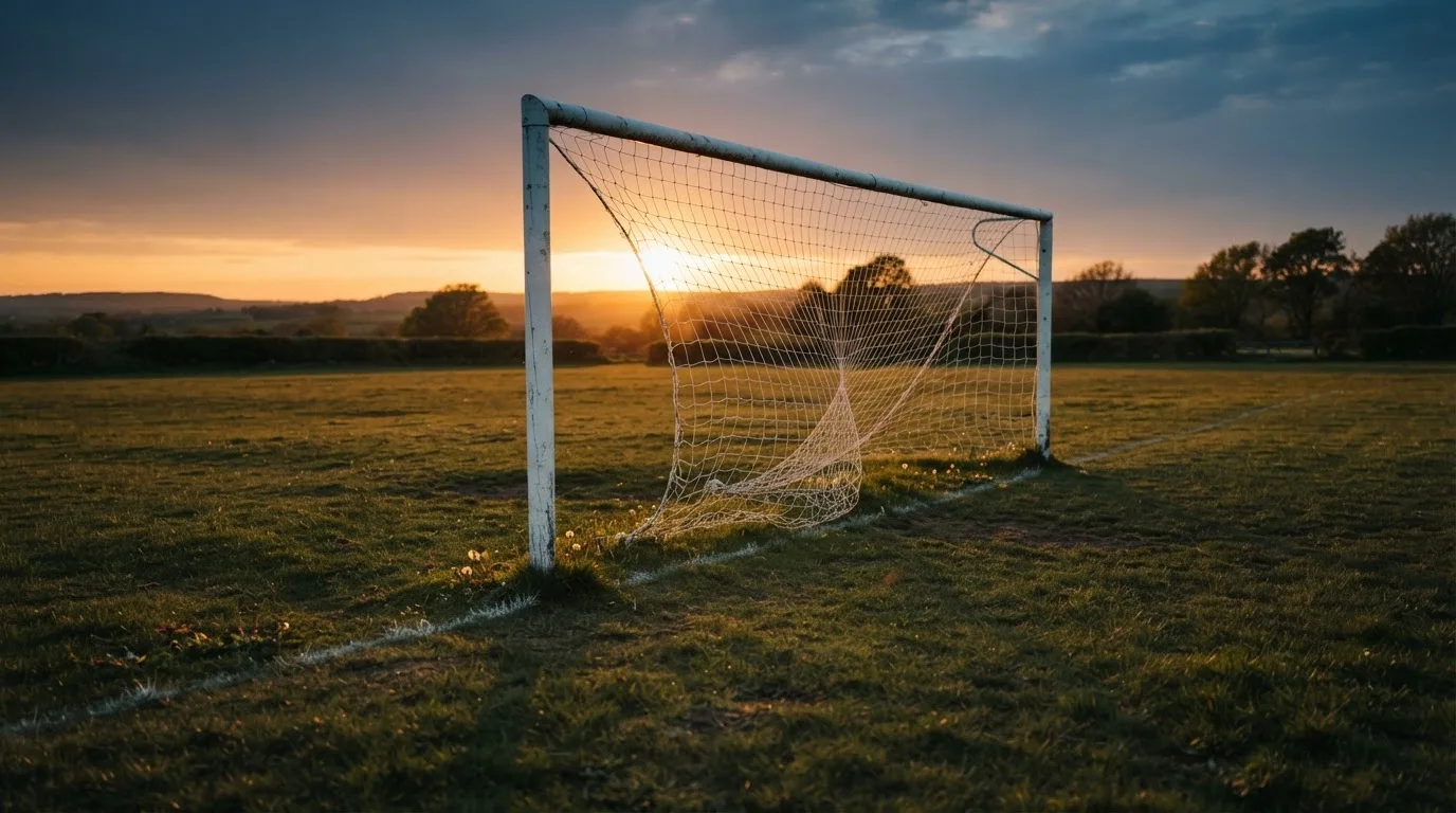 Ein Fußballtor mit Netz bei Sonnenuntergang auf einem leeren Platz, die Schatten der Torpfosten liegen lang auf dem Gras.