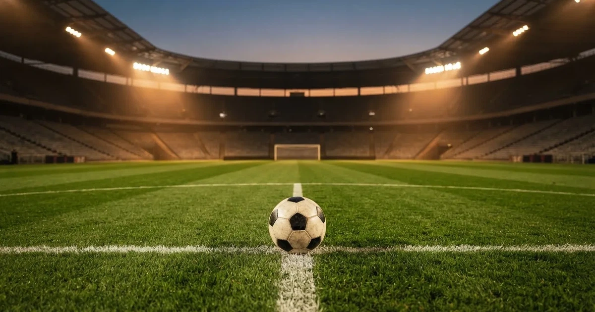 Ein Fußball liegt auf dem Anstoßpunkt eines leeren Rasenplatzes, im Hintergrund eine große Stadiontribüne im Abendlicht.