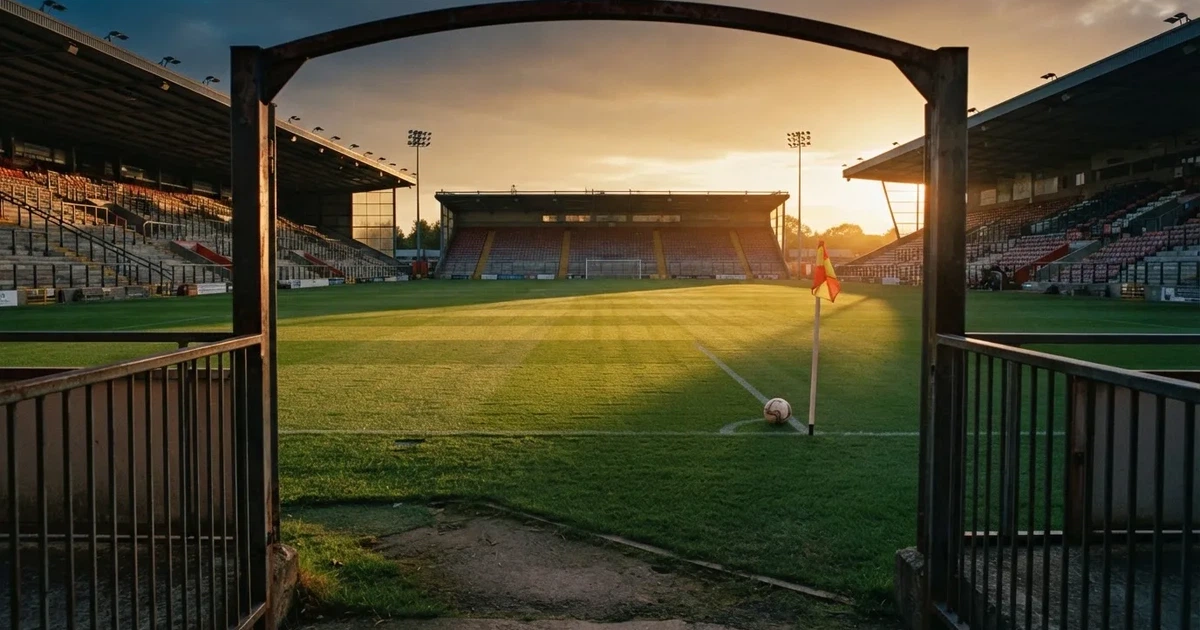 Blick durch ein Stadiontor auf einen grünen Rasen im Abendlicht, ein Ball ruht an der Eckfahne, die Tribünen sind leer.
