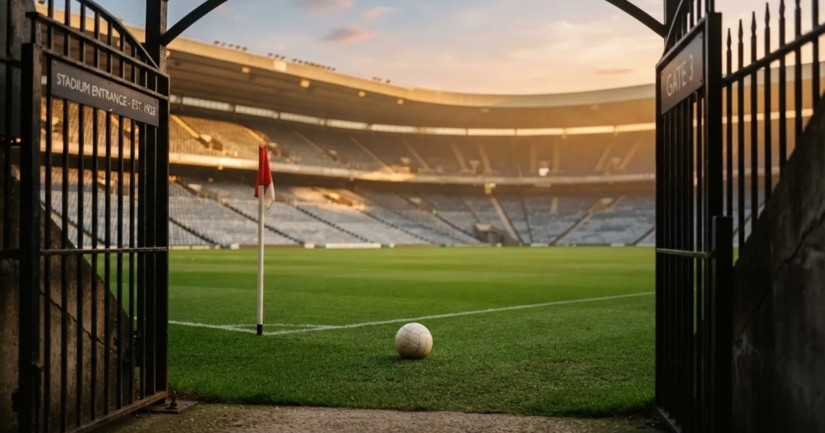 Blick durch ein Stadiontor auf einen grünen Rasen im Abendlicht, ein Ball liegt an der Eckfahne, die Ränge sind leer.