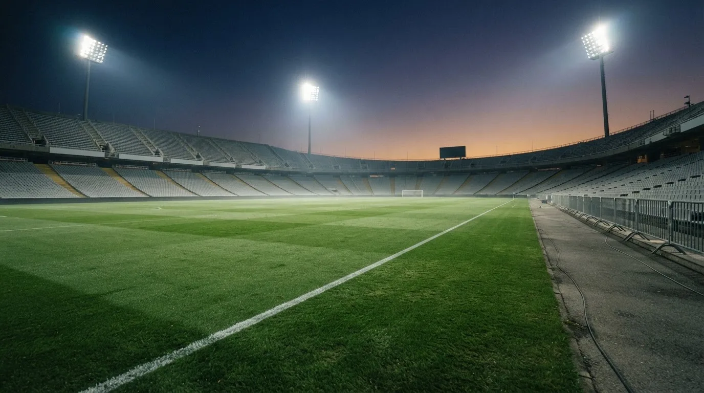 Eine leere Stadiontribüne unter Flutlicht aus der Ferne betrachtet, die Rasenfläche leuchtet grün in der Dämmerung.