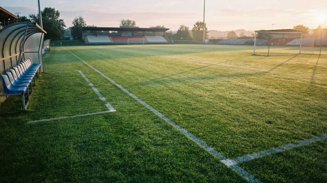 Ein Fußballplatz mit frisch gezogenen Kreidelinien im ersten Morgenlicht, leere Ersatzbänke am Spielfeldrand.