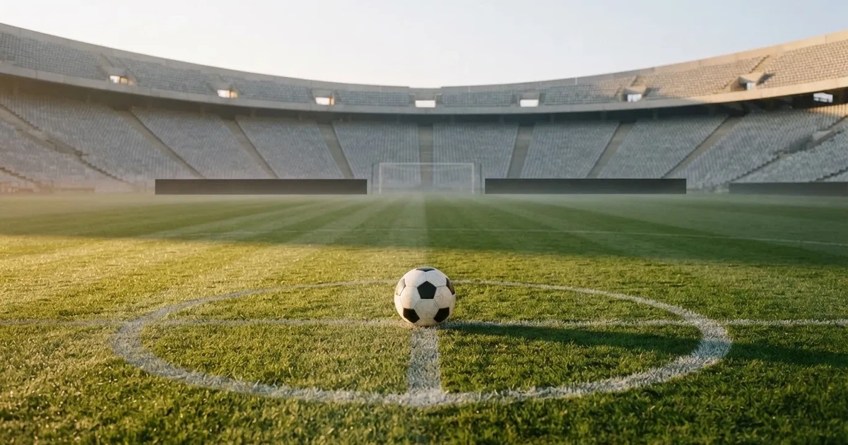 Ein Fußball liegt auf dem Mittelkreis eines Stadionrasens am frühen Morgen, im Hintergrund leere Ränge ohne erkennbare Beschriftung.
