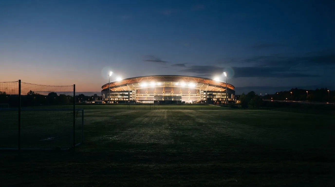 Ein Stadion unter Flutlicht aus der Ferne, davor ein stilles Feld im Dämmerlicht.