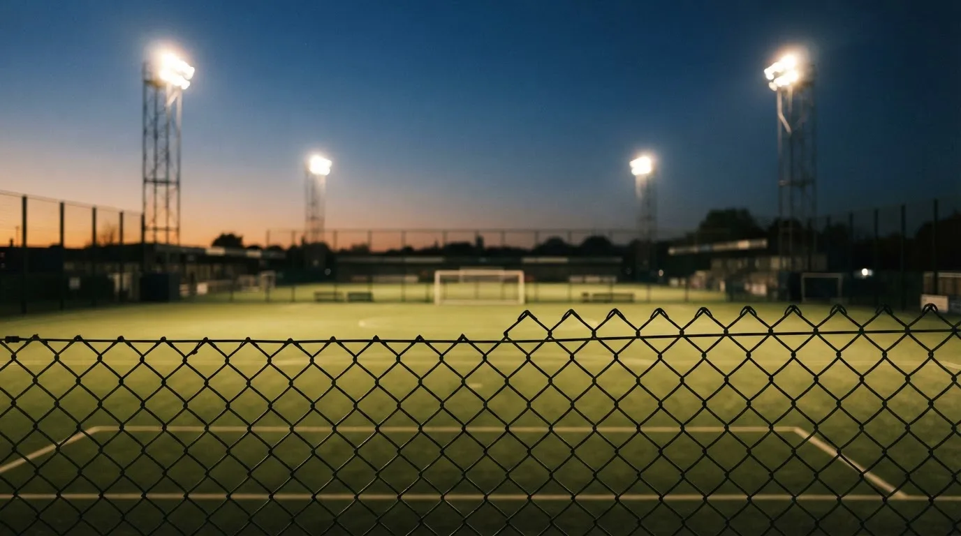 Eine Flutlichtanlage über einem leeren Sportplatz am Abend, im Vordergrund ein Maschendrahtzaun.