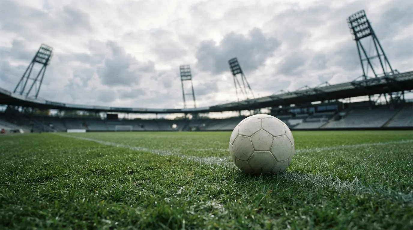 Ein Fußball auf einem Rasenplatz, im Hintergrund Flutlichtmasten und ein bewölkter Himmel.