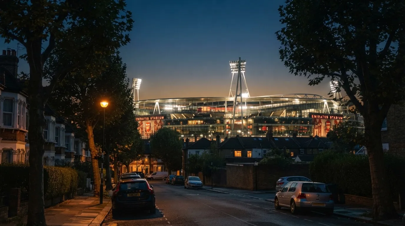 Ein Stadion aus der Ferne bei Flutlicht, davor eine ruhige Straße in der Abenddämmerung.