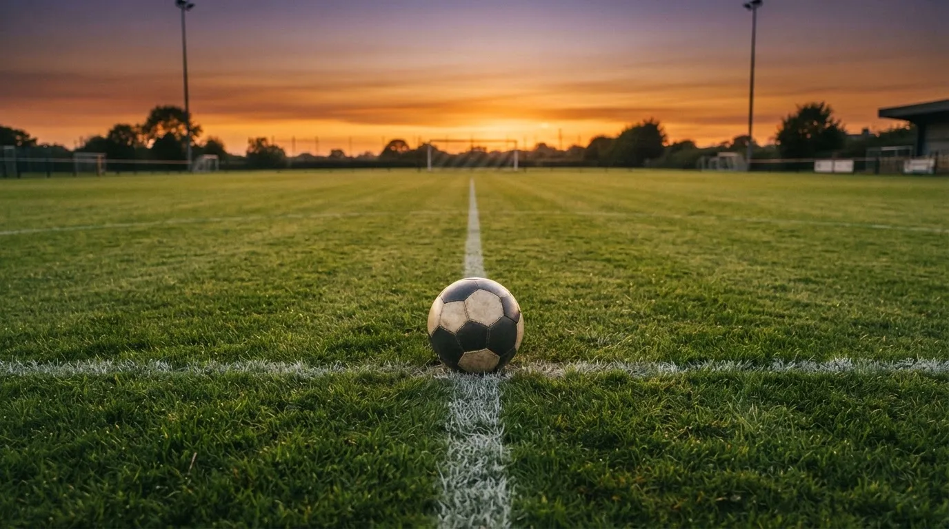 Ein Fußball liegt in der Mitte eines leeren Rasenplatzes, am Horizont färbt sich der Himmel orange.