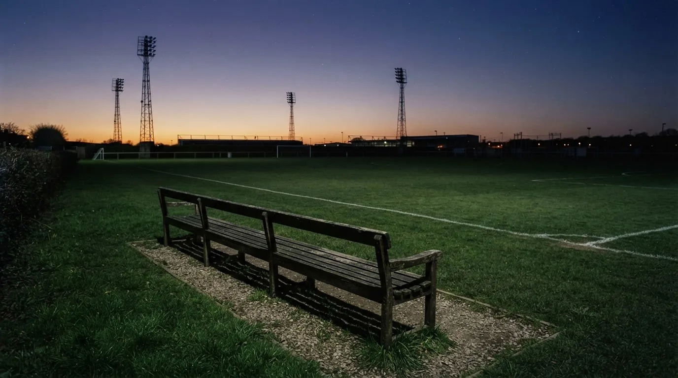 Eine leere Parkbank am Rand eines Fußballfelds in der Abenddämmerung, im Hintergrund ragen dunkle Flutlichtmasten in den Himmel.