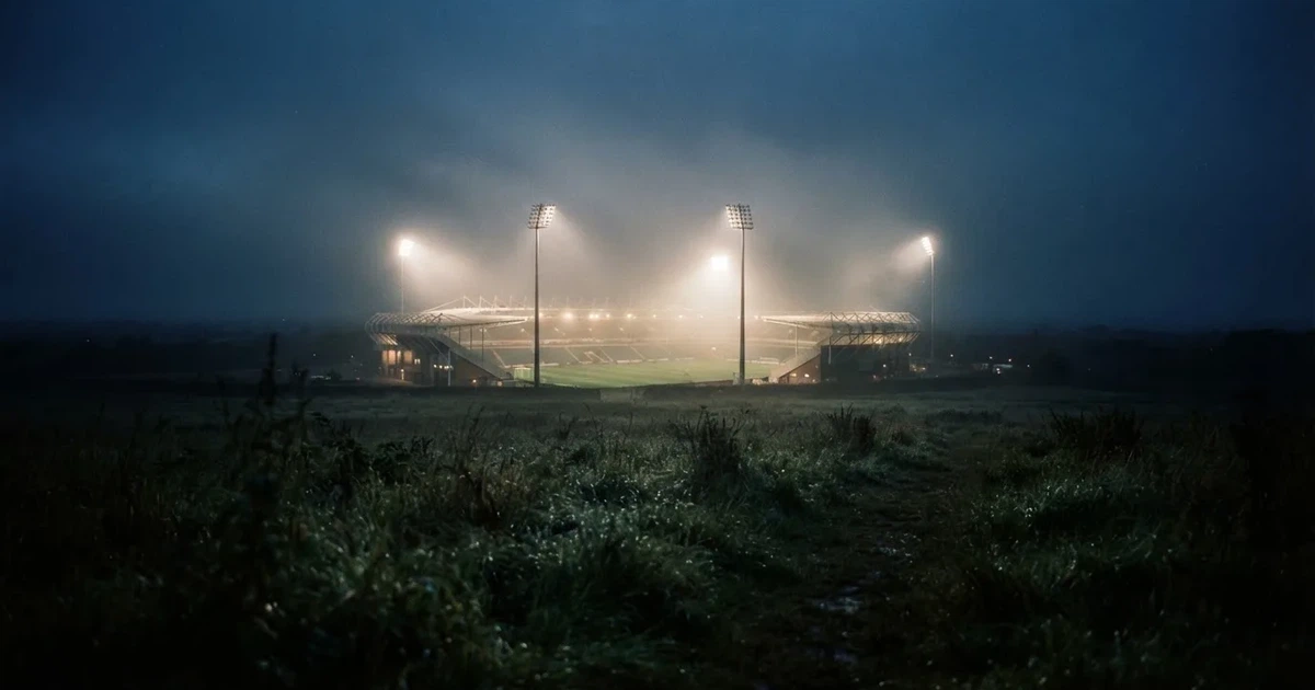 Ein Stadion unter Flutlicht, aus großer Entfernung über eine dunkle Wiese fotografiert, am Himmel zieht Nebel auf.