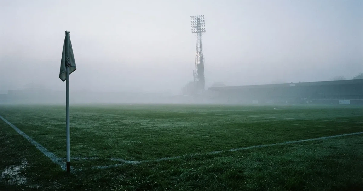 Eine Eckfahne steht im Nebel am Rand eines leeren Rasenplatzes in der Morgendämmerung, dahinter ein Flutlichtmast.