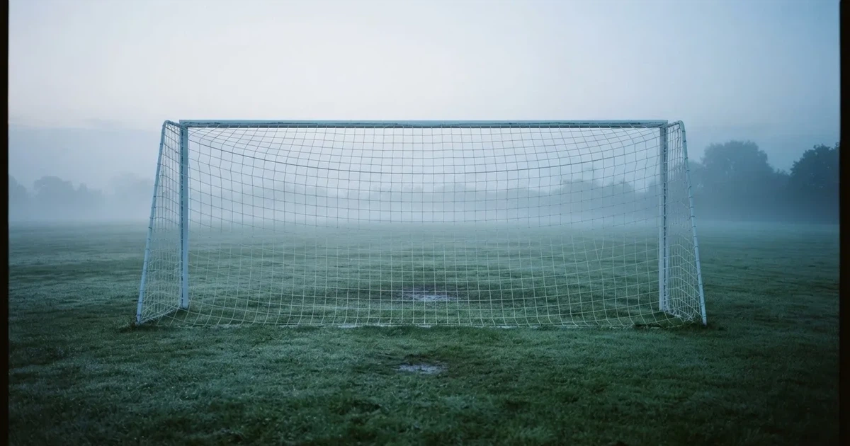 Ein Fußballtor mit Netz steht auf einem leeren Platz im Nebel der Morgendämmerung, der Rasen ist taufeucht.