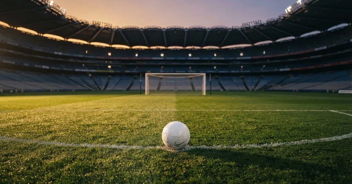 Ein Fußballplatz mit frisch gemähtem Rasen im Abendlicht, ein Ball liegt im Strafraum, im Hintergrund unscharf eine leere Tribüne.