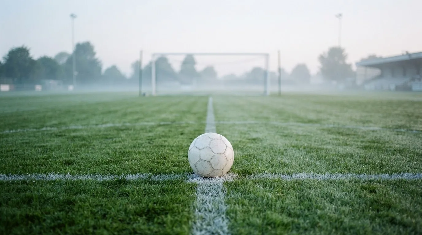 Ein Fußball liegt auf dem Elfmeterpunkt eines leeren Rasenplatzes, im Hintergrund unscharf ein Tor im Morgendunst.