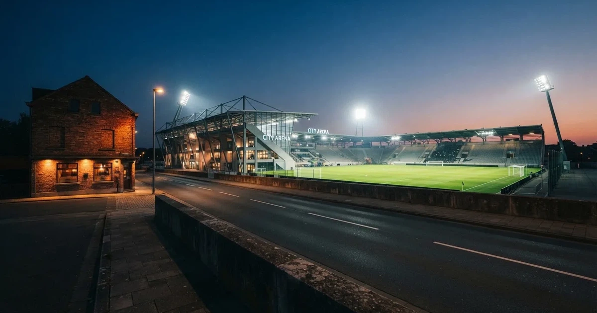 Ein Fußballstadion bei Flutlicht aus der Ferne gesehen, davor eine Straße und leuchtende Fenster eines Nebengebäudes.