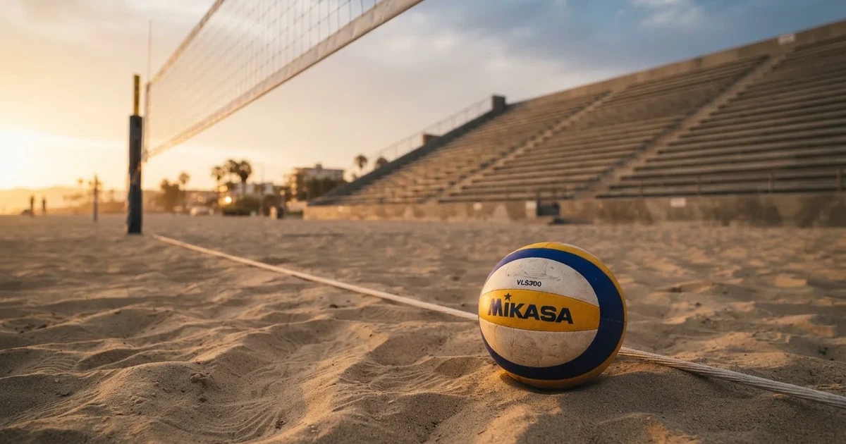 Ein Volleyball liegt auf dem Sand eines Beachvolleyball-Feldes neben dem gespannten Netz, im Hintergrund eine leere Tribüne bei Abendlicht.