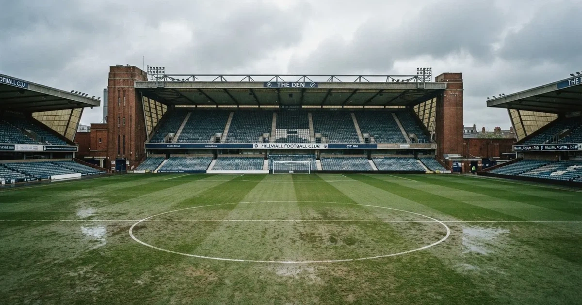 Ein englisches Fußballstadion mit rotbraunen Backsteintribünen unter bedecktem Himmel, davor ein nasser Rasen mit Spielfeldlinien.