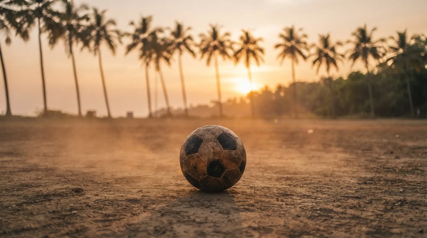 Ein Fußball liegt auf einem staubigen Bolzplatz mit Palmen im Hintergrund in warmem Abendlicht.