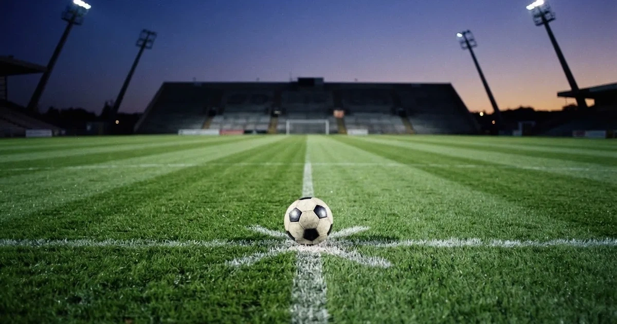 Ein Fußball liegt auf dem Anstoßpunkt eines leeren Rasenplatzes, im Hintergrund eine Tribüne und Flutlichtmasten in der Abenddämmerung.