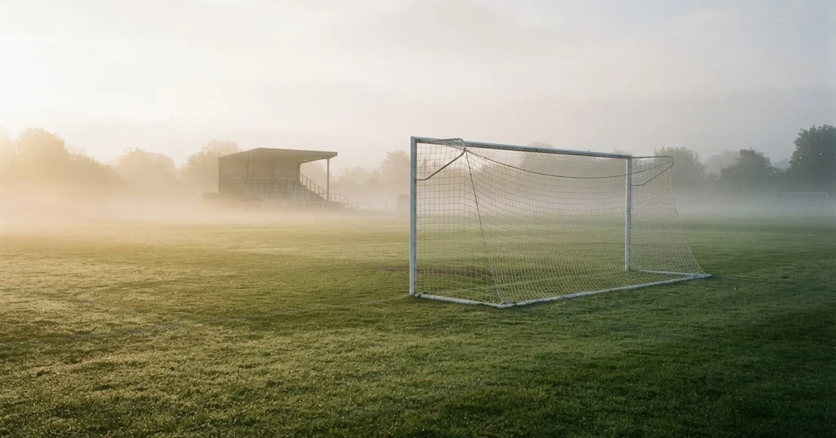 Ein Fußballtor mit Netz steht im Morgennebel auf einem leeren Rasenplatz, im Hintergrund die Silhouette einer kleinen Tribüne.