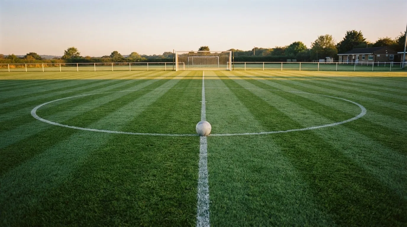 Ein Fußball liegt allein auf dem Anstoßpunkt eines leeren Rasenplatzes im Morgenlicht, am Rand ein niedriger Zaun.