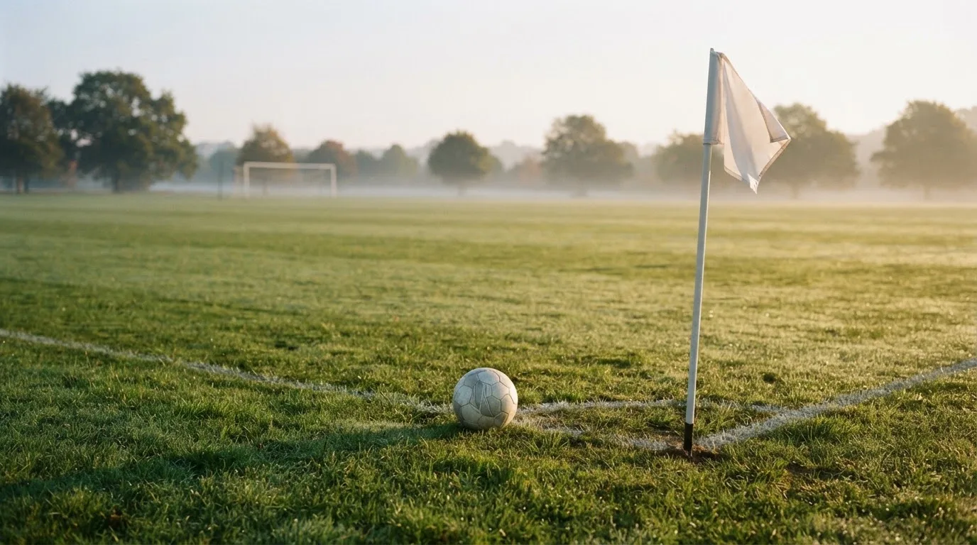 Ein Fußball liegt auf Rasen neben einer Eckfahne ohne Aufdruck auf einem leeren Platz im Morgenlicht.