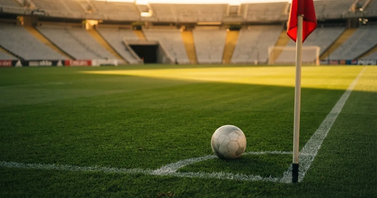 Ein Fußball liegt am Viertelkreis an der Eckfahne eines Rasenplatzes, im Hintergrund leere Tribünen in warmem Abendlicht.