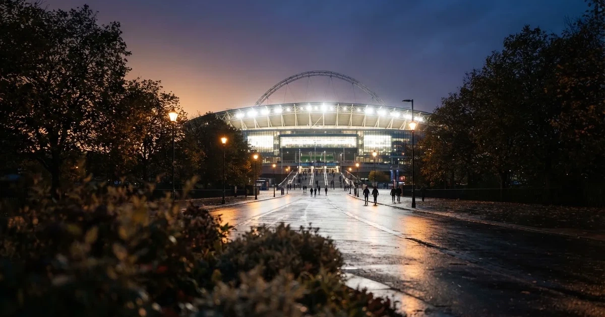 Ein großes Fußballstadion mit hell erleuchtetem Rund, aus der Ferne am Abend gesehen, davor eine breite Allee mit Laternen.