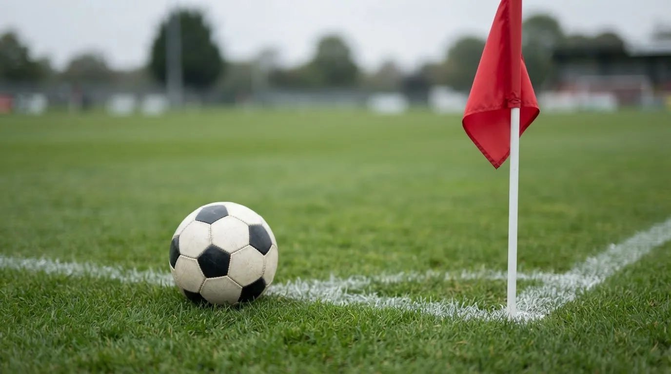 Ein Fußball liegt neben einer Eckfahne auf kurz gemähtem Rasen unter bewölktem Himmel.