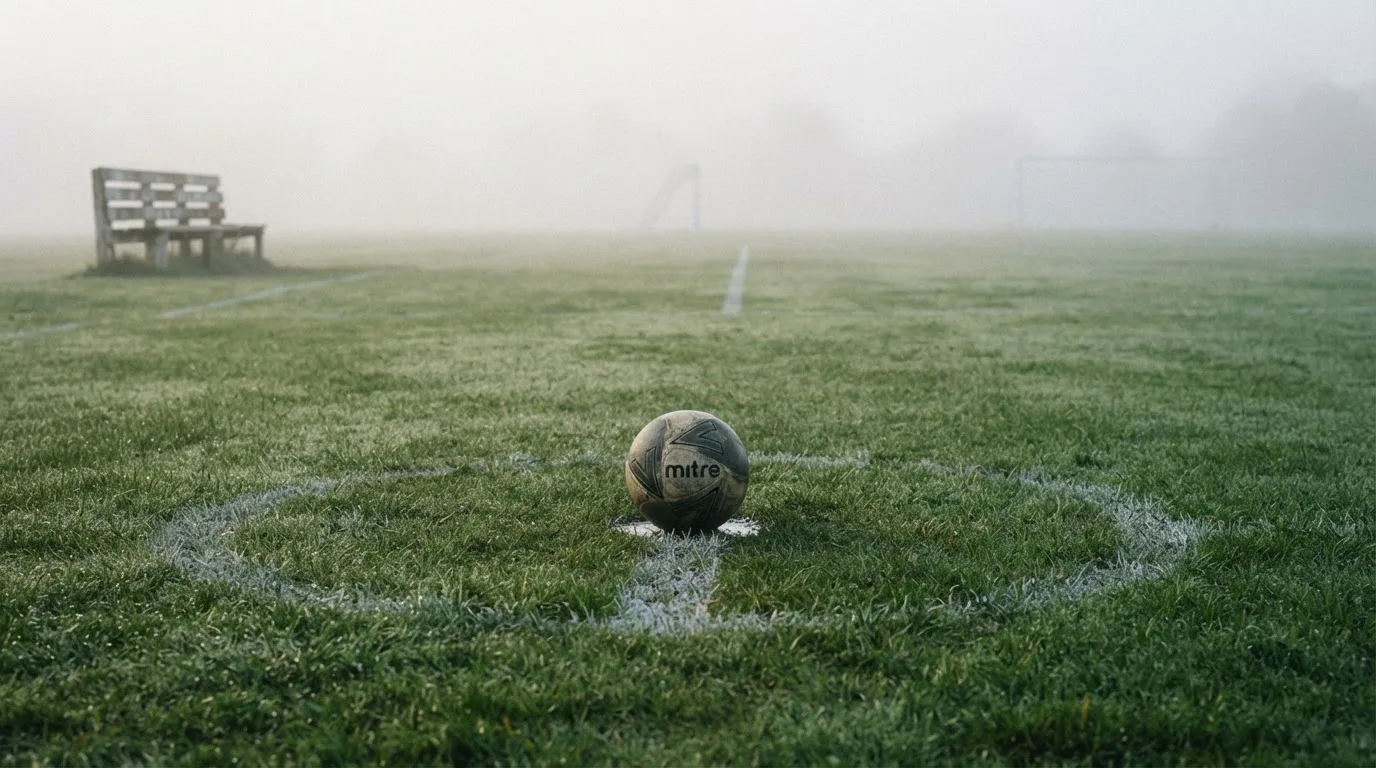Ein Fußball liegt in der Mitte eines leeren Rasenplatzes im Morgennebel, am Rand steht eine einzelne Auswechselbank.
